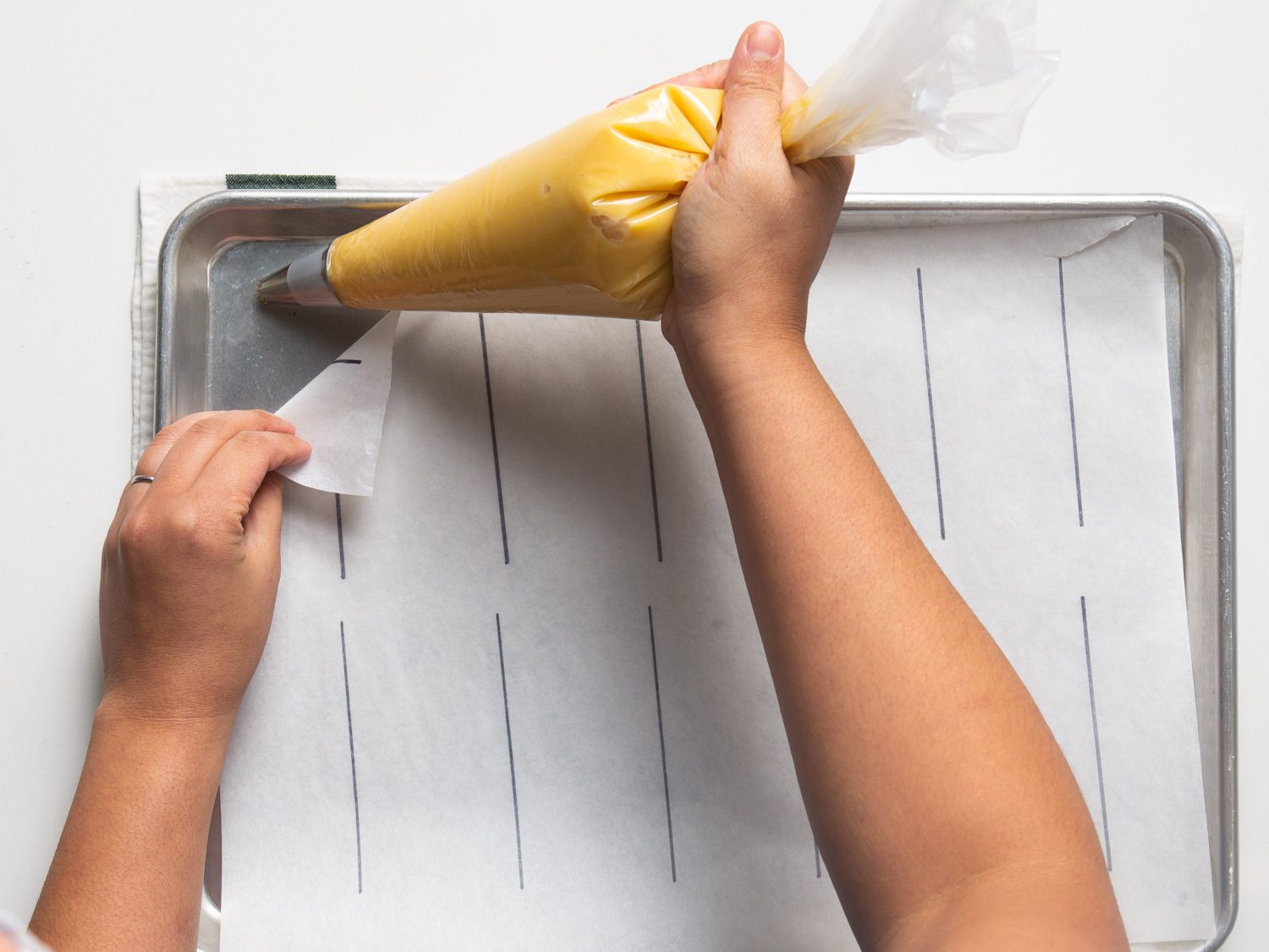 Choux paste being squeezed under all four corners of the parchment paper to prevent it from moving. 