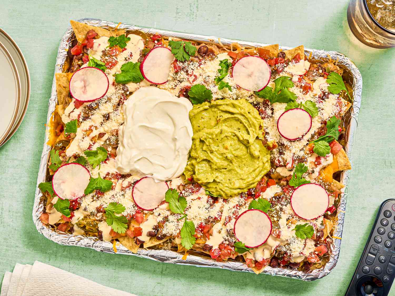 Loaded nachos with a tv remote, plates, napkins and a drink on the edges of the frame, on a light green surface