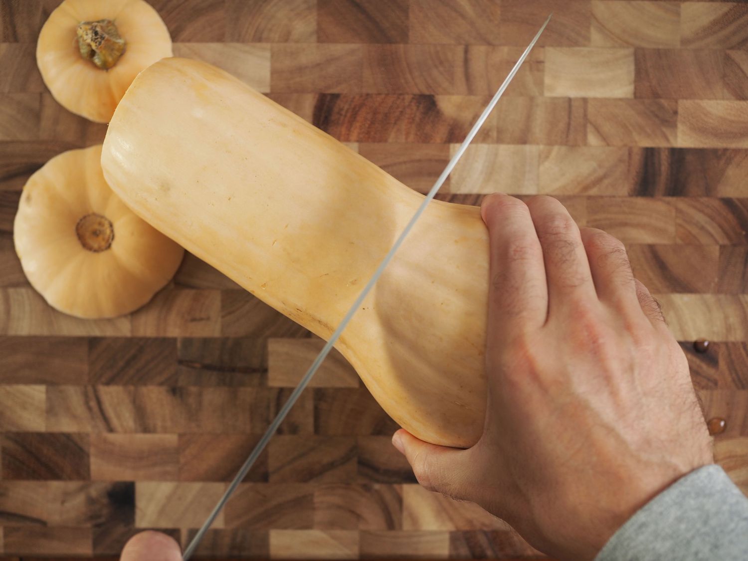 A hand firmly gripping a butternut squash, with a knife slicing off its neck.