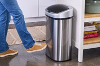 A person uses their foot to open the lid on the iTouchless 13-Gallon Oval Stainless Steel Sensor Trash Can