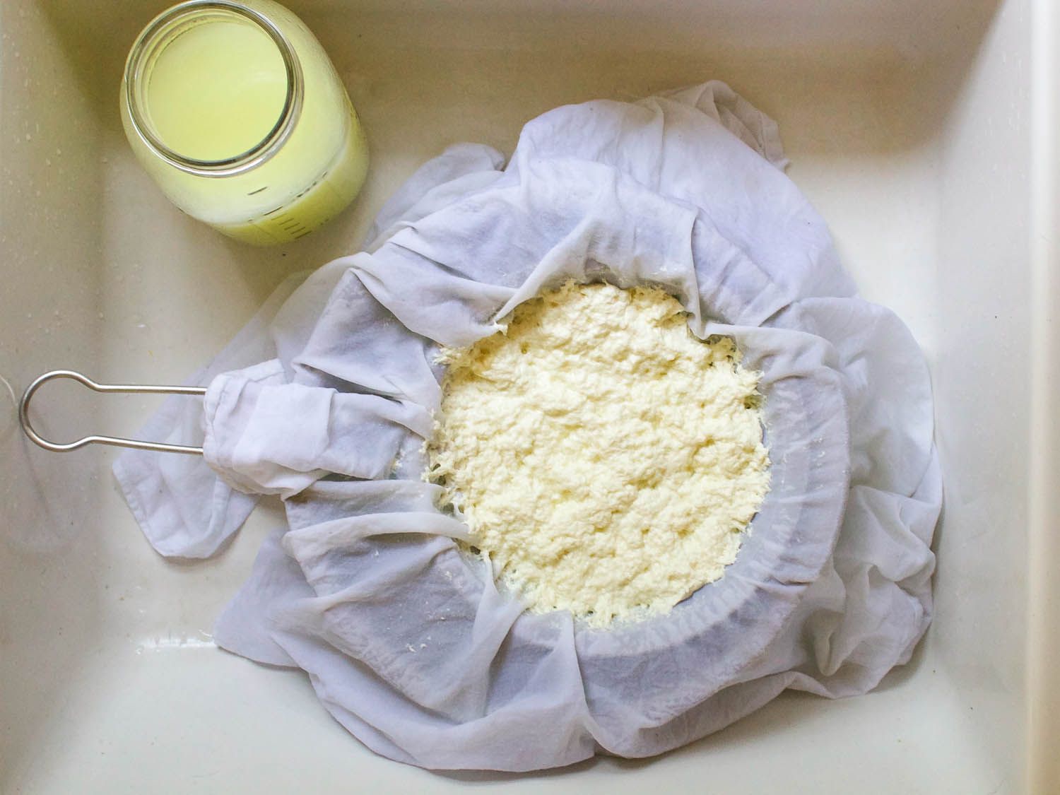 Curds draining in a cheesecloth-lined colander, a jar of whey nearby.