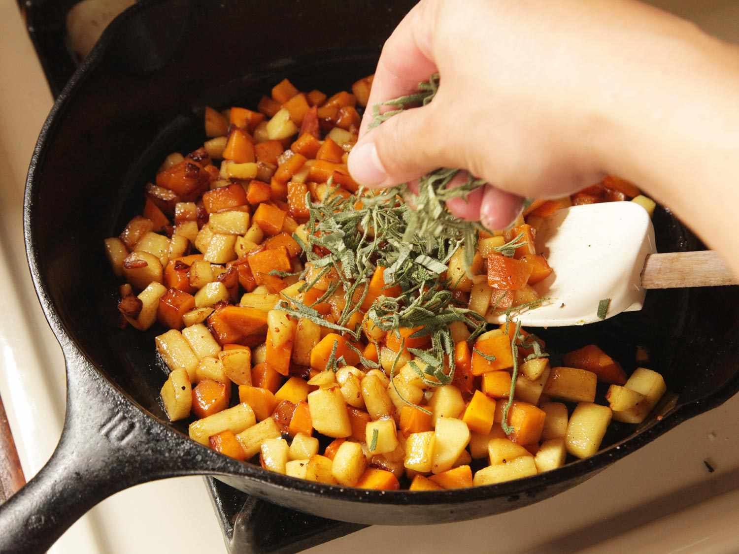 Sprinkling minced sage on a pan containing sauteed squash cubes and apple cubes.