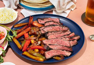 Platter of grilled skirt steak fajitas, peppers, and onions, with tortillas, cheese, and other toppings in the background.