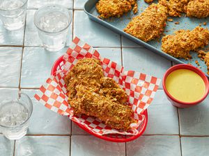 Honey mustard chicken tenders in a basket with dip, more tenders on a tray in the background.
