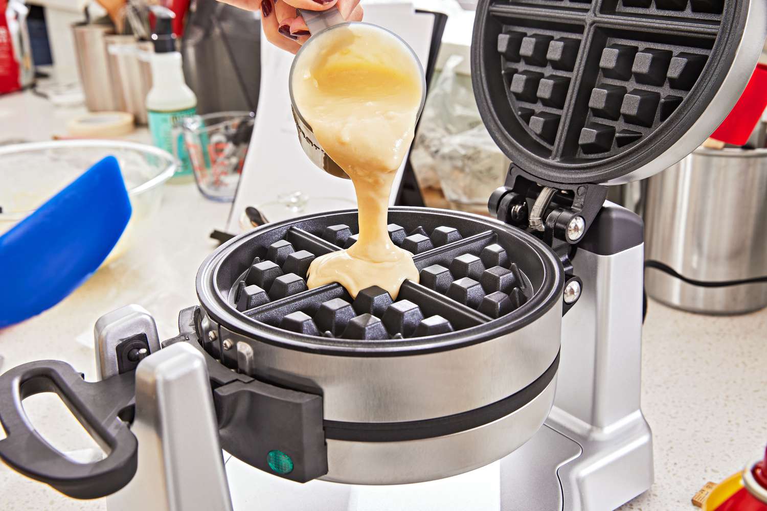 A hand pouring waffle batter onto a waffle iron with its lid open