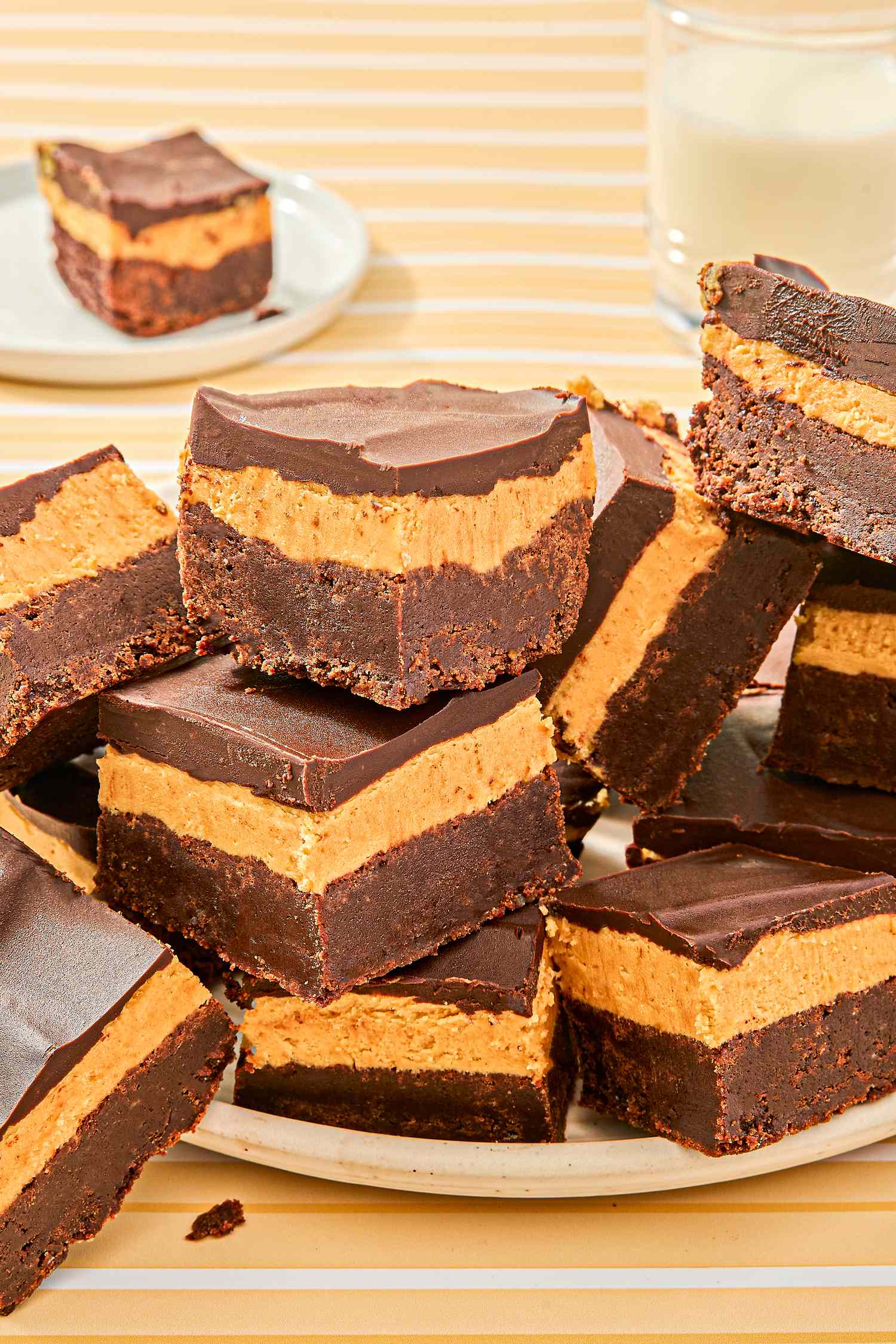 Stacked brownies with a peanut butter layer and chocolate topping on a plate with a glass of milk and a plate in the background