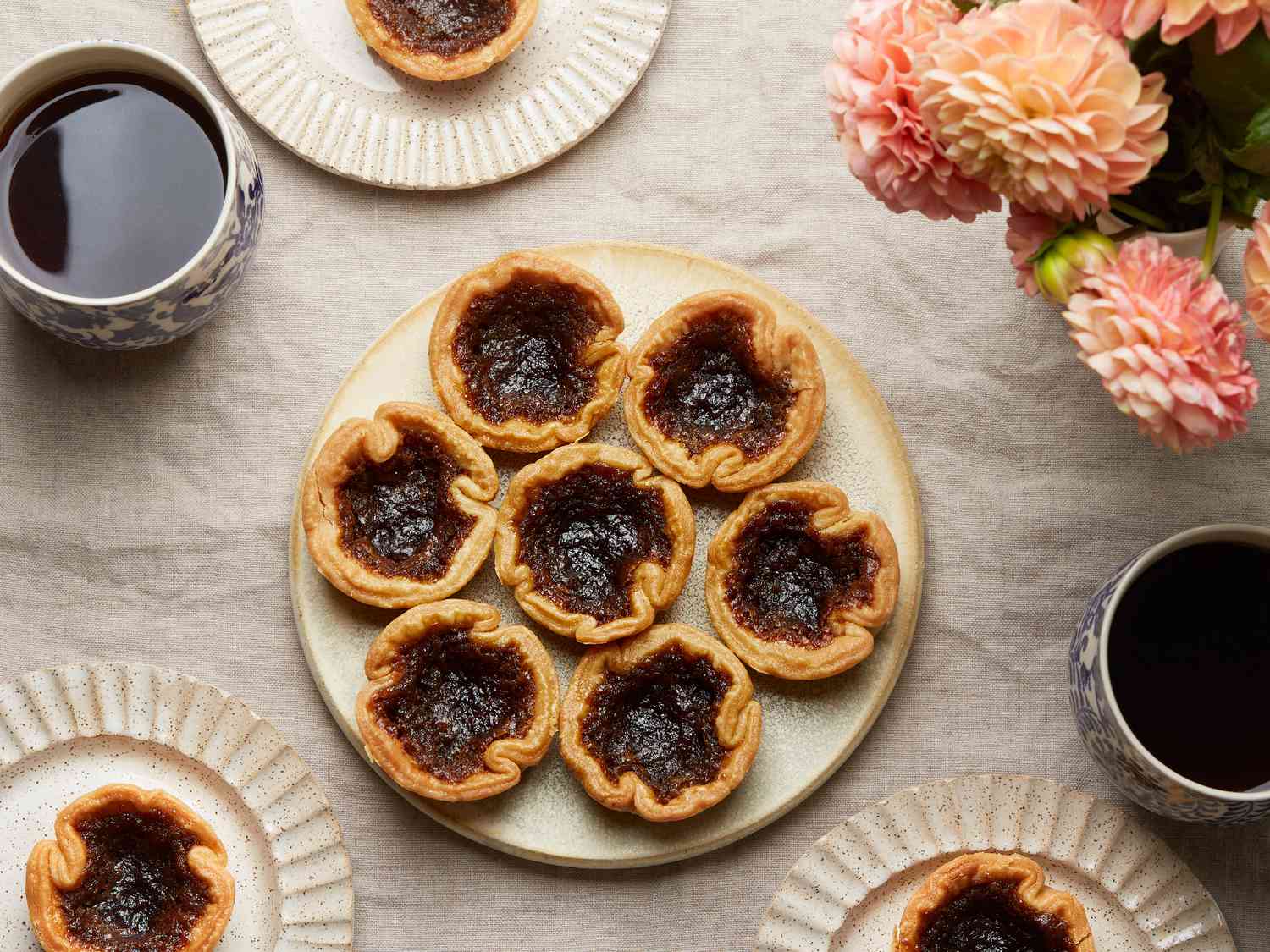 Overhead of platter with Canadian butter tarts. 3 smaller plates have one tart each. Coffee and flowers on the tabletop, with linen tablecloth.