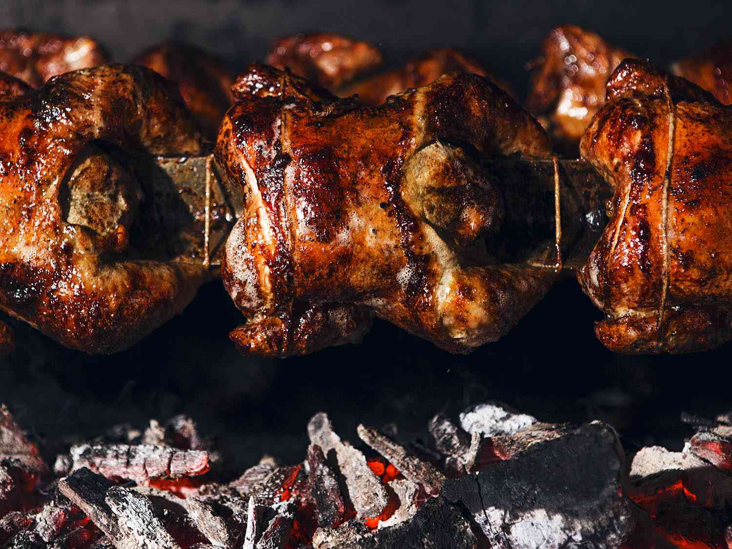 Three whole chickens roasting on a spit over coals.