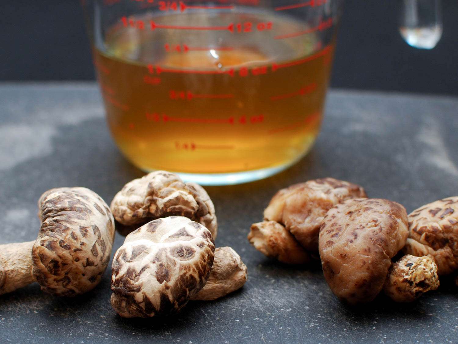 Several rehydrated shiitake mushrooms in front of a cup of their golden-brown soaking liquid.