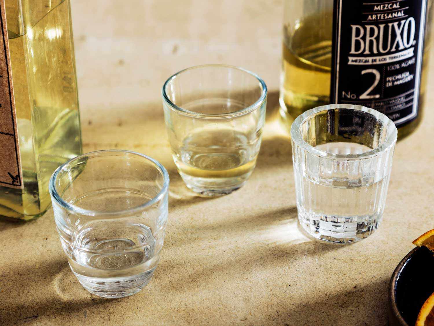 Three tasting cups half filled with mezcal next to 2 bottles of mezcal.