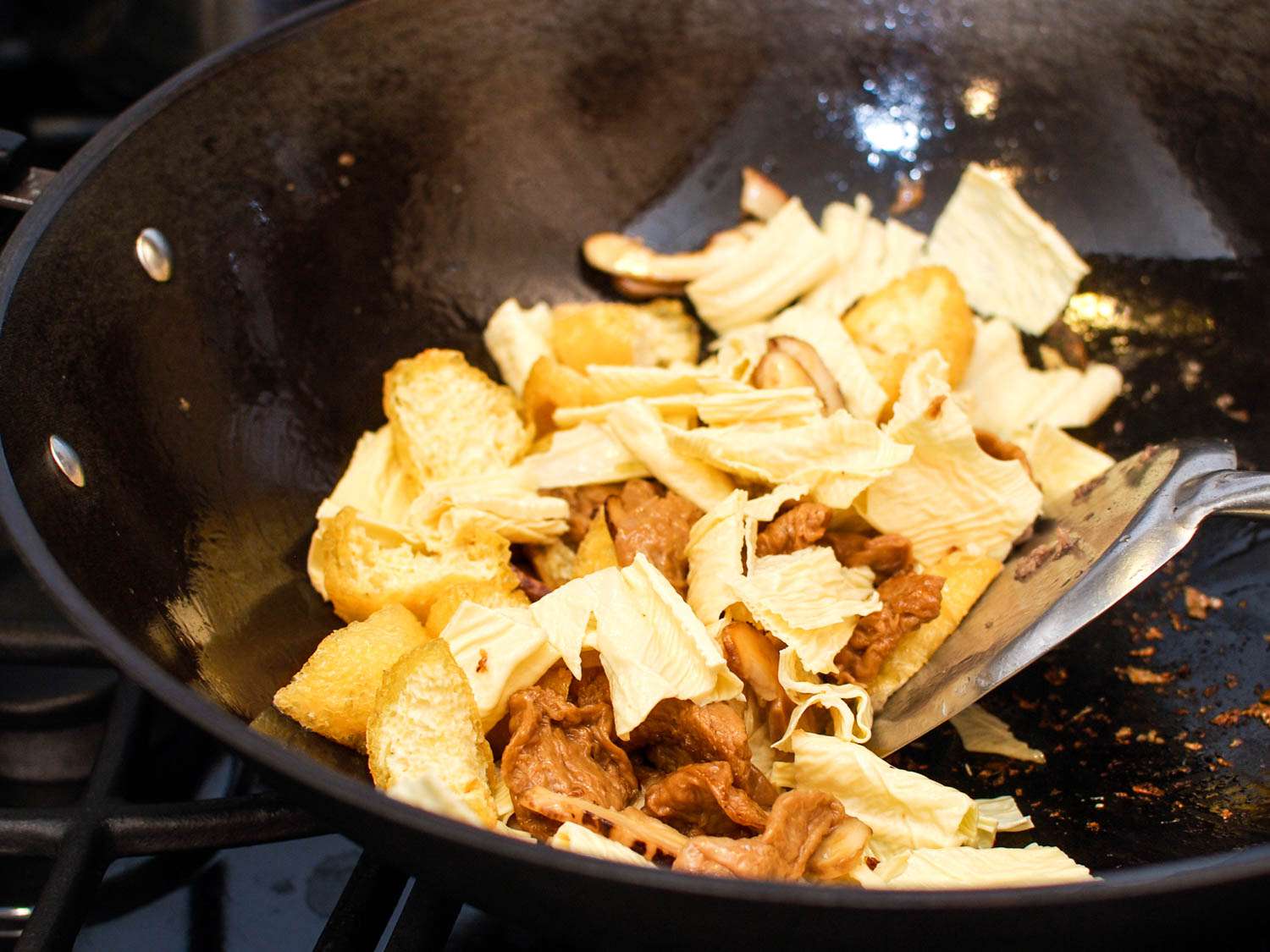 Stirring chai pow yu, mushrooms, tofu puffs, and bean curd sticks in a wok with a metal spatula.