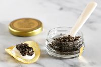 An open jar of Olma beluga hybrid caviar next to a potato chip topped with crème fraîche and a scoop of caviar