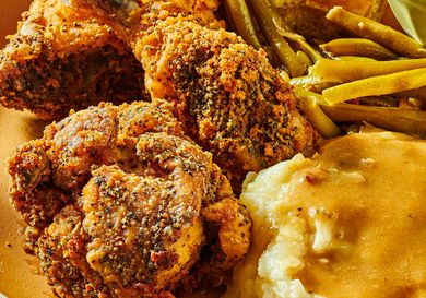 Dish of fried chicken with coleslaw, greenbeans, and mashed potatoes, with a fork and serving dishes in the background