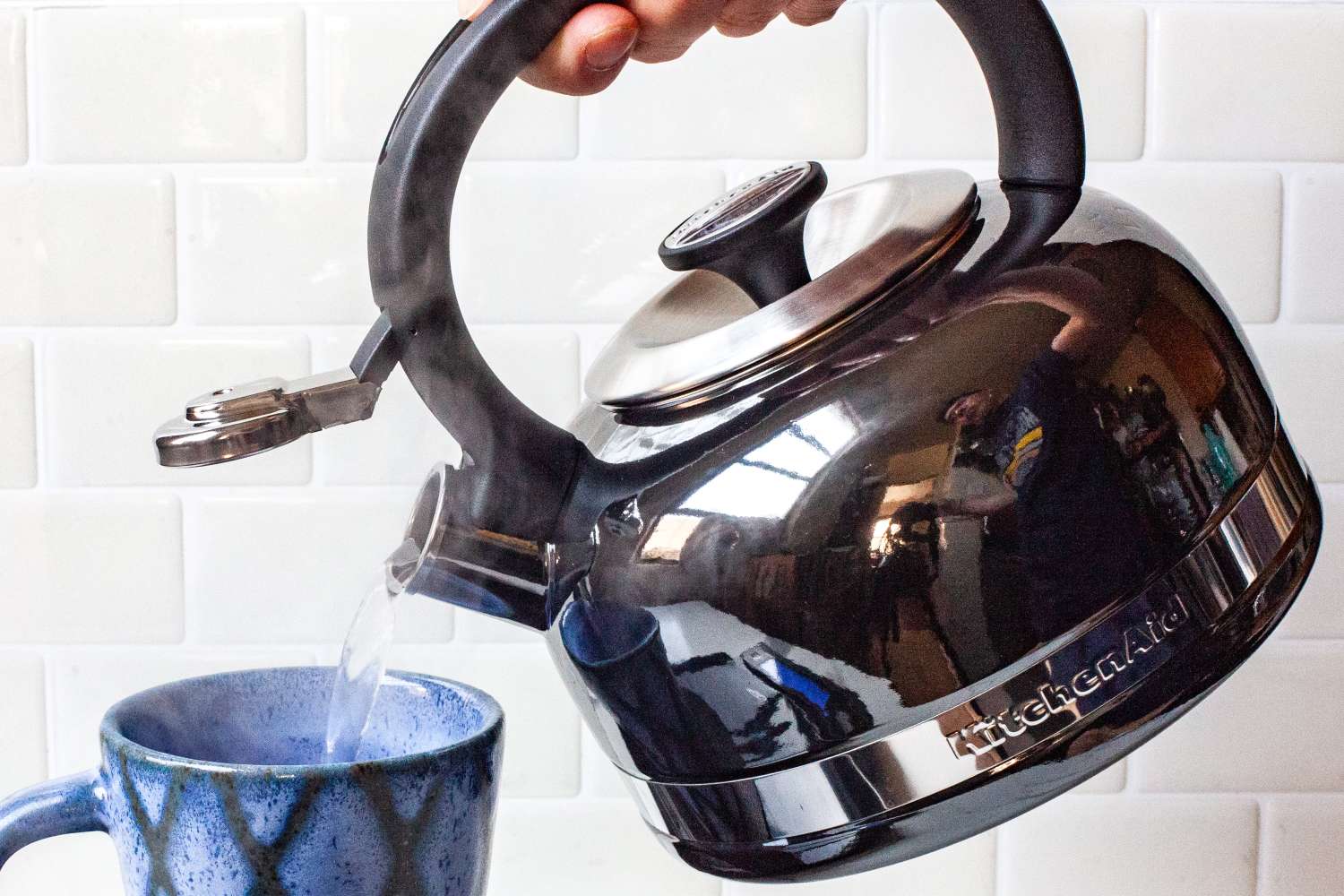 A hand pouring hot water from a kettle into a mug on a tiled backsplash background