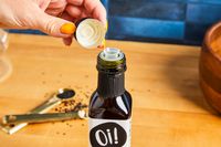 A hand opening a small bottle labeled "Oi!" with measuring spoons filled with ingredients in the background