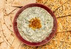 A plate of creamy spinach dip garnished with fried onions surrounded by flatbread and sesame crackers