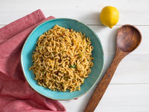 A bowl of pasta salad with Spanish chorizo and piquillo peppers. A lemon, wooden spoon, and red kitchen towel are next to the bowl.