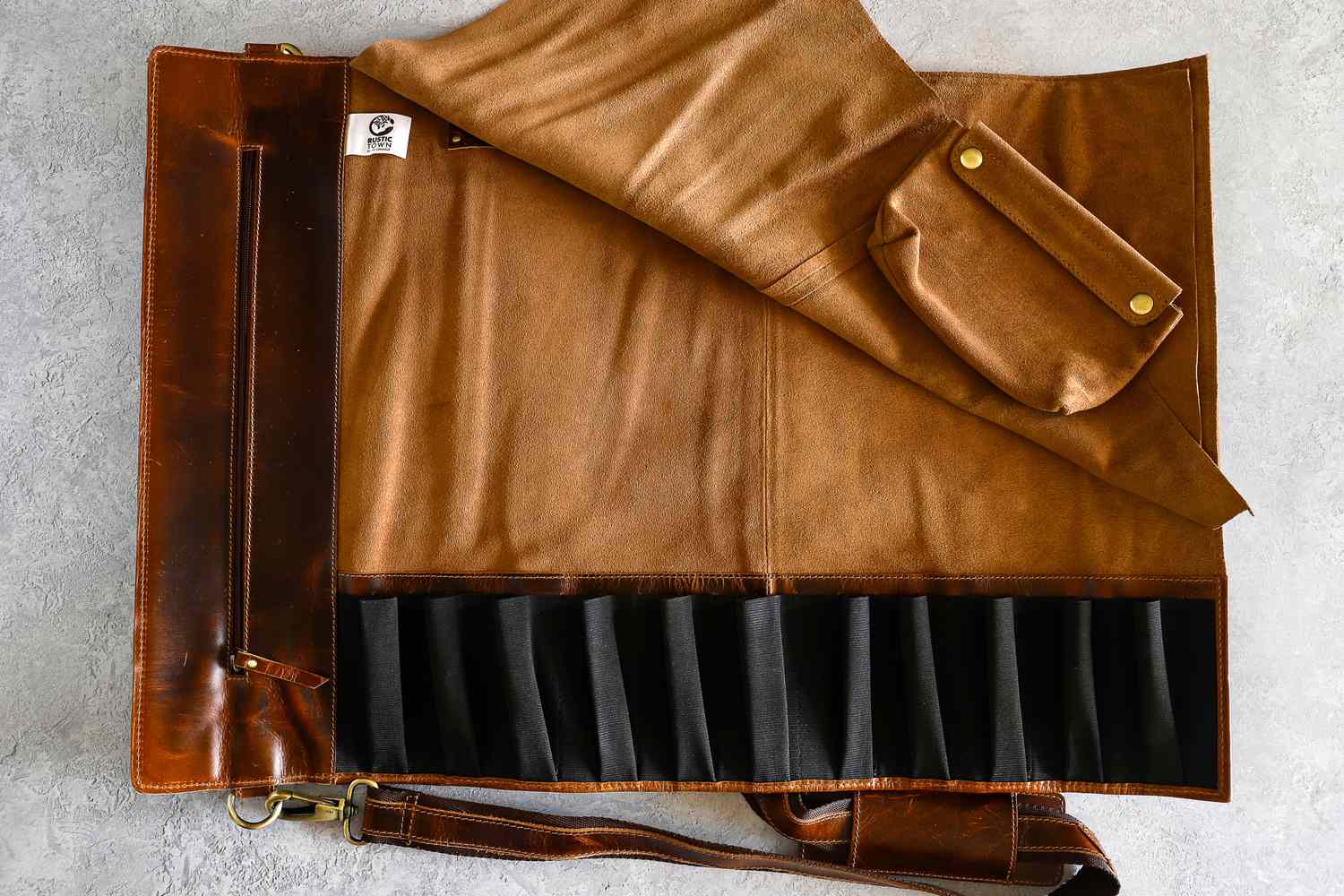 An opened Rustic Town Genuine Leather Chef Knife Roll with compartments a strap and a small pouch on a textured surface