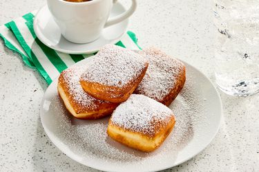 Beignets on a white plate with powdered sugar, with a coffee, green and white striped napkin, and a marble surface
