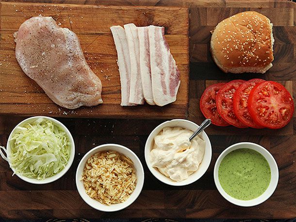An overhead shot of all the components of the sandwich arranged on a work surface: pounded and seasoned raw chicken breast, raw bacon slices, sesame bun, tomato slices, jalapeño-avocado sauce, mayonnaise, crushed potato chips, shredded lettuce.