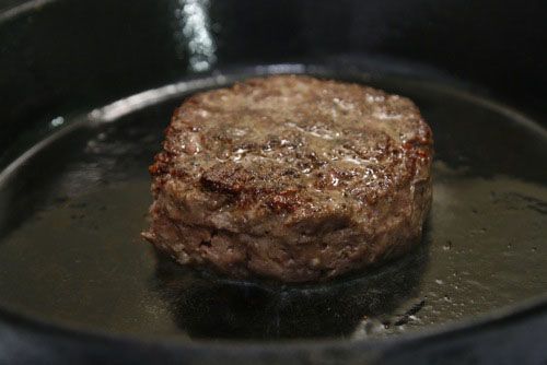 A thick, reverse-seared hamburger in a cast iron pan. The burger is browned and glistening.