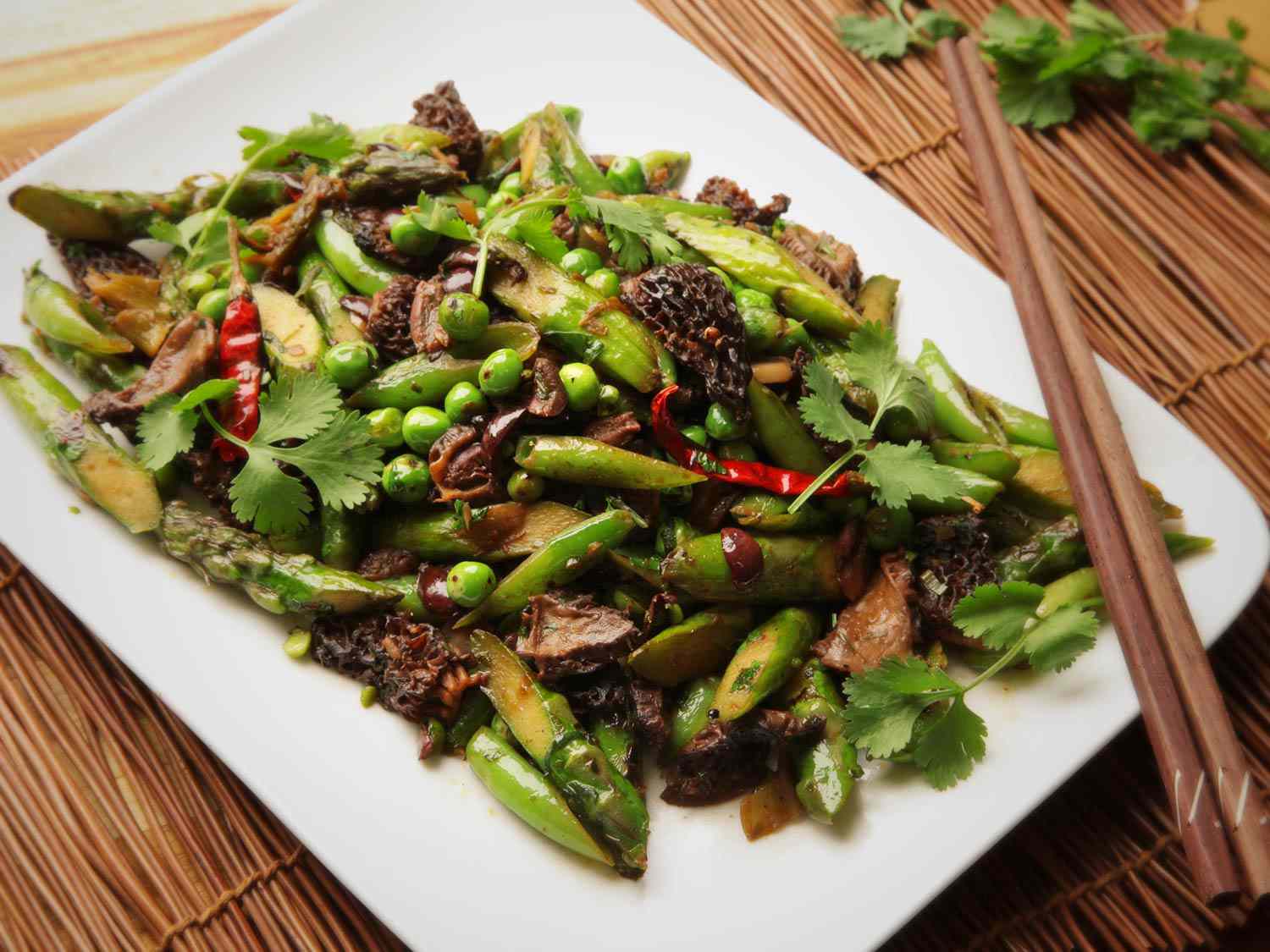 Stir-fried asparagus, English peas, snap peas, morel mushrooms, and black olives on a white plate
