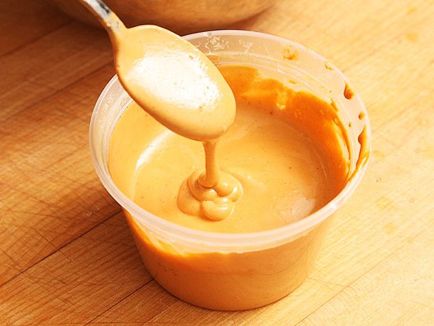 Vegan cheese sauce in a pint-sized deli container is spooned out and held overhead, showing its thick, pourable consistency as it drips back nito the container.