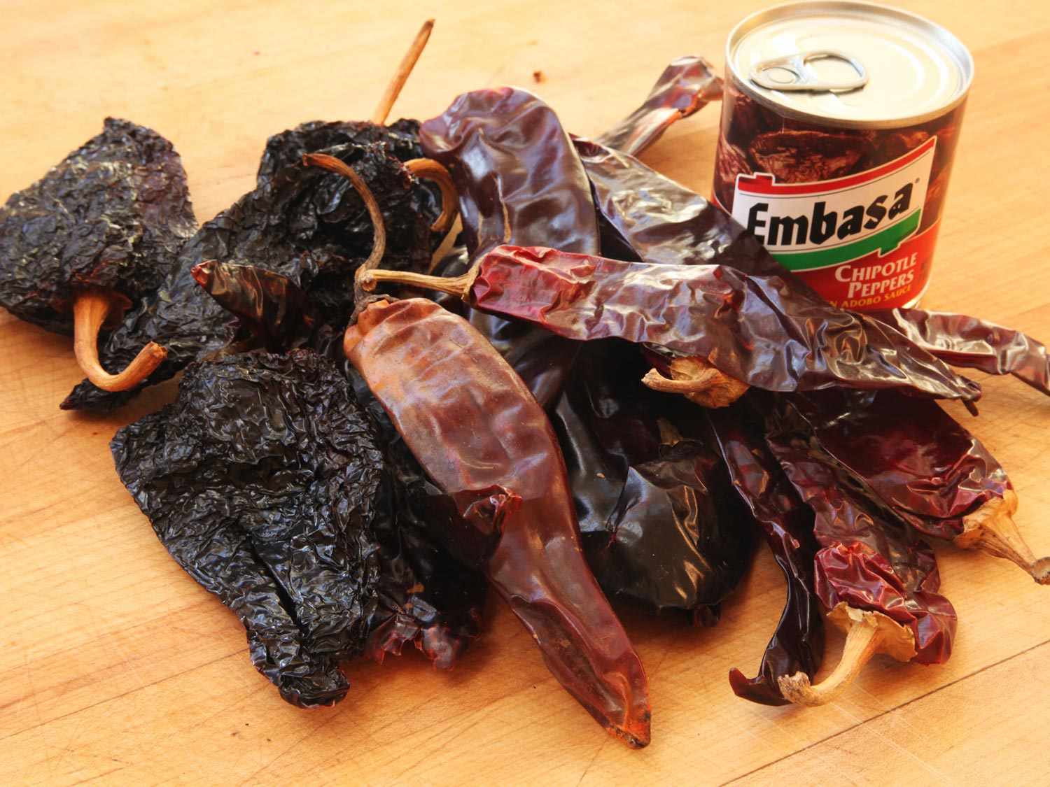 Dried guajillo, pasilla, and ancho chiles, plus a can of chipotles en adobo, on a wooden cutting board for carne asada marinade.