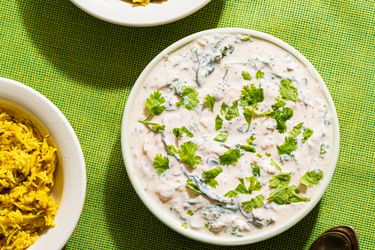 A bowl of raita garnished with herbs next to two bowls of spiced rice and a spoon