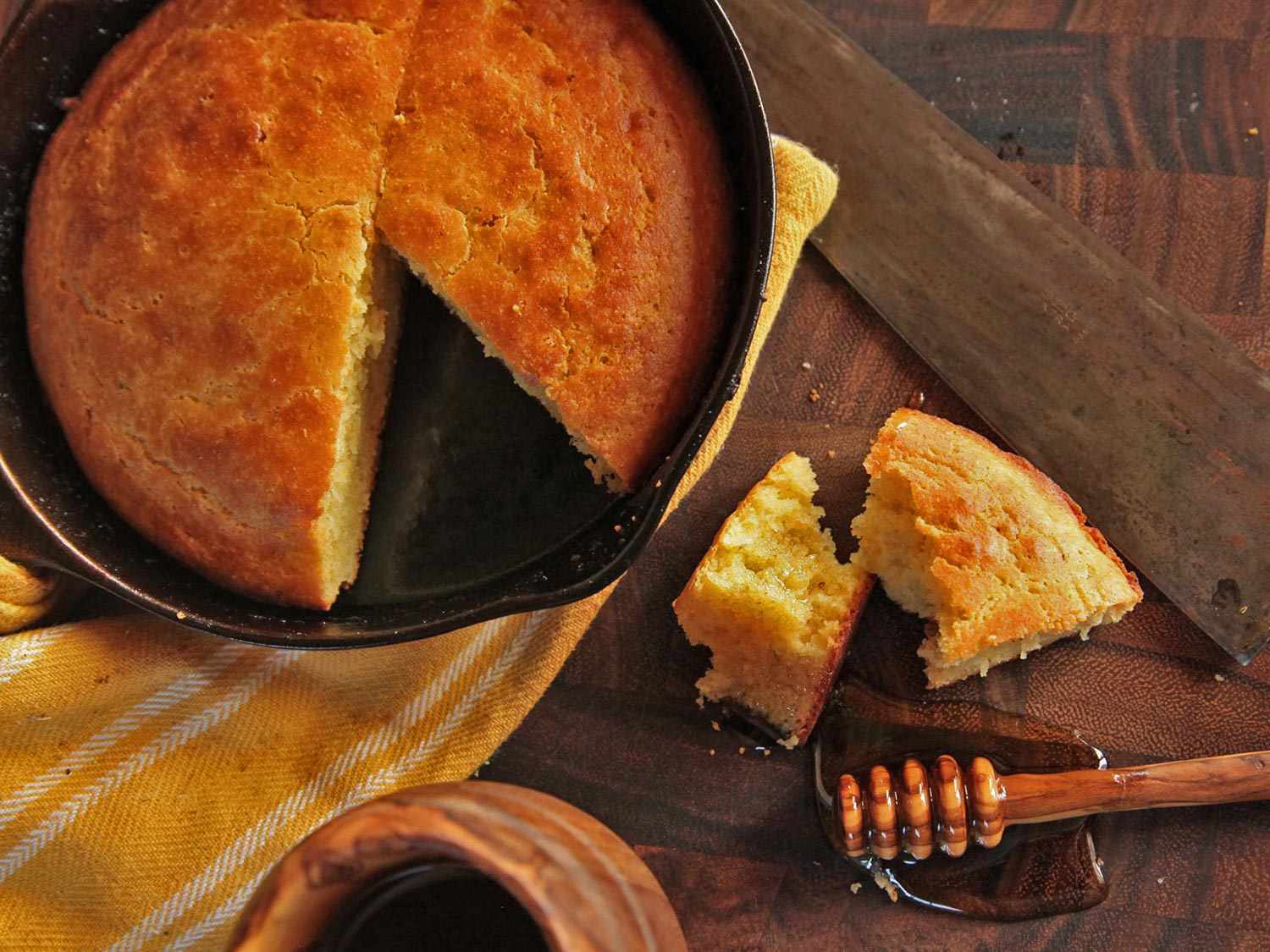 Overhead view of a cast iron skillet full of brown butter cornbread. A wedge has been cut, broken into chunks, and placed next to a puddle of honey.