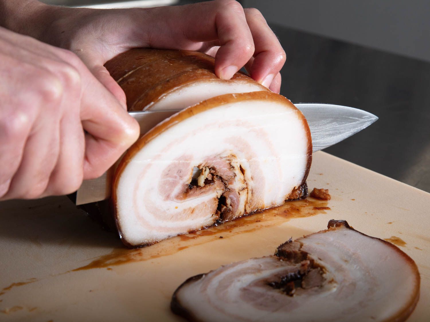 Hand cutting a slice of cooked rolled up pork belly with a chef's knife.