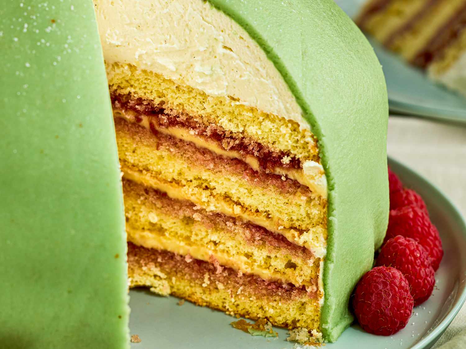 A crosssection view of a layered Princess cake with green marzipan covering sponge cake layers and raspberry filling garnished with raspberries