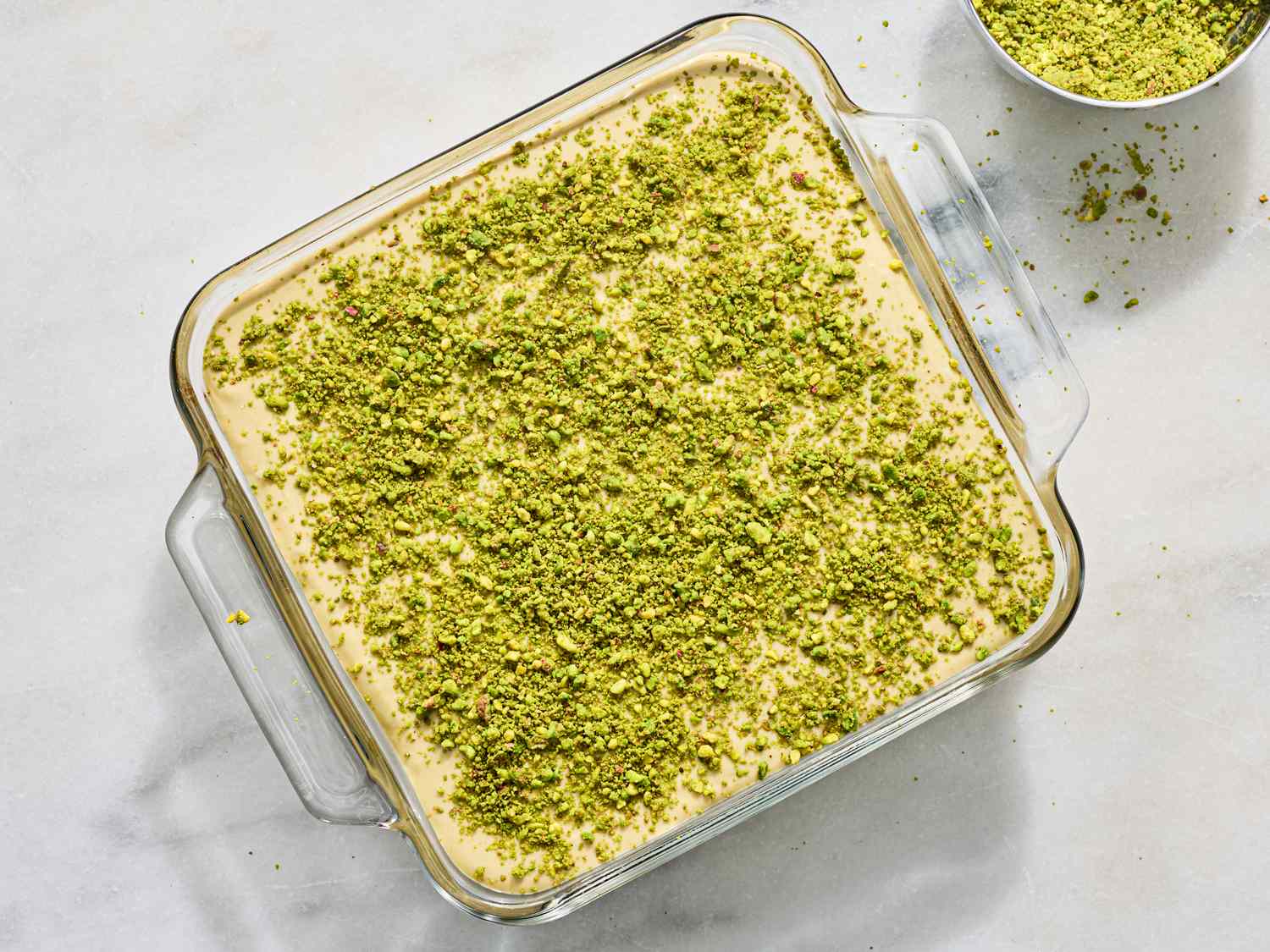 A dish of pistachio tiramisu in a square glass container on a light surface topped with crushed pistachios with a bowl of pistachios nearby