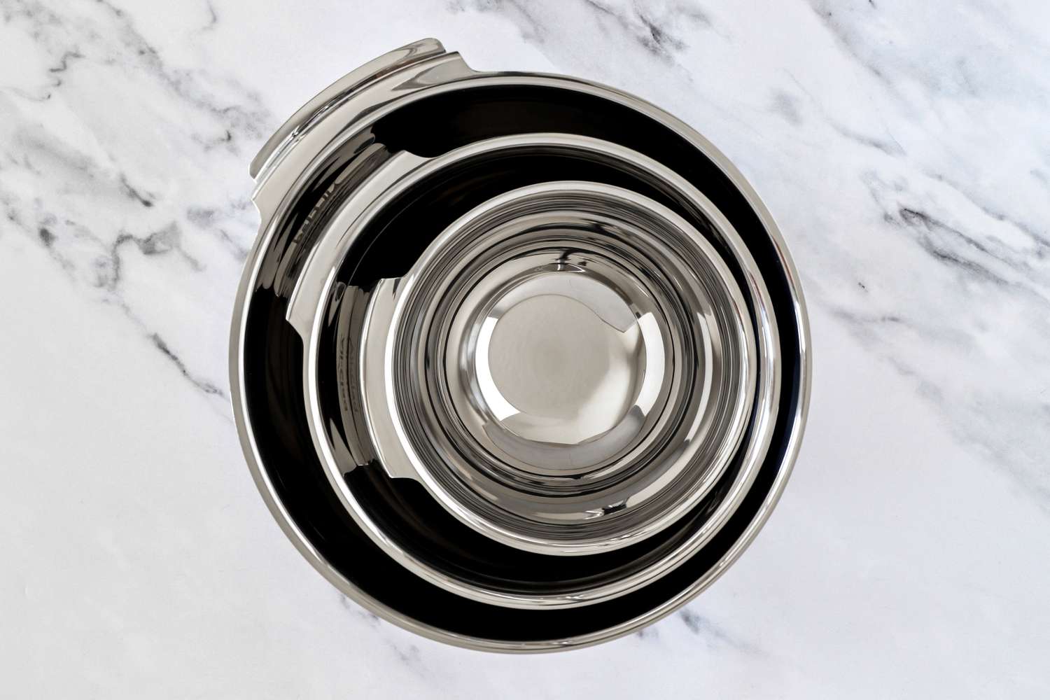 A set of stainless All-Clad mixing bowls stacked together placed on a marble surface