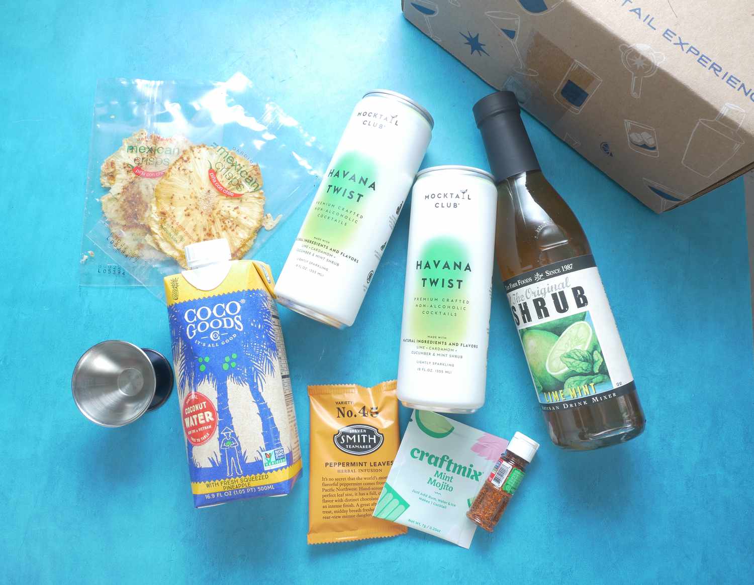 A few cans of drinks, a bottle of shrub, and a few packets of various ingredients (tea, tajin) on a blue surface