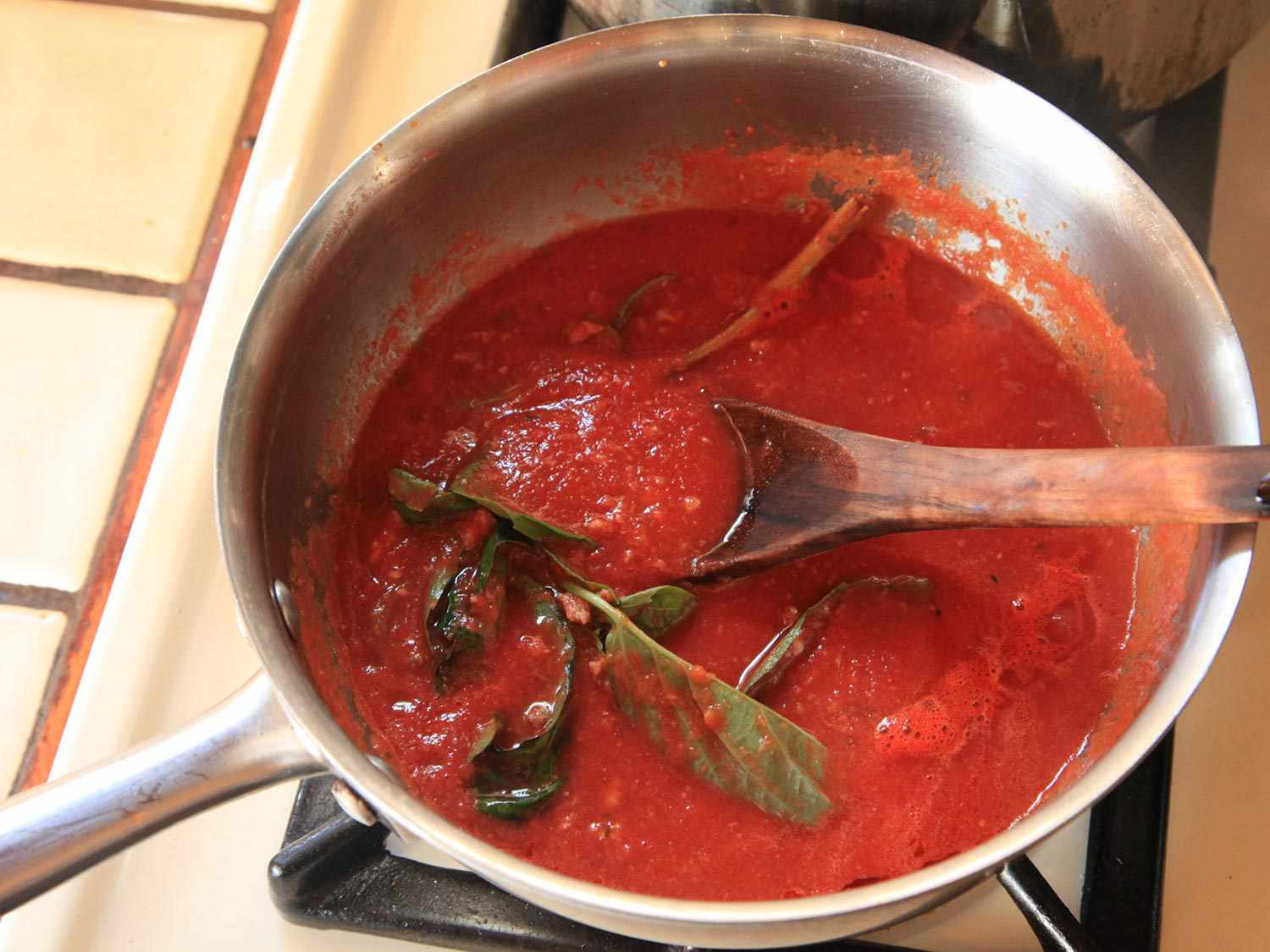 A pot of canned tomatoes and basil sprigs cooking on the stovetop, with a wooden spoon in the sauce.