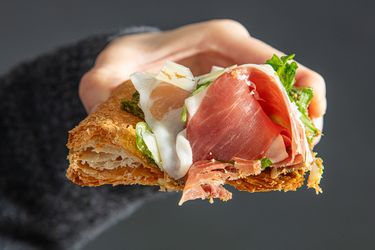Hand holding a piece of flaky pastry topped with cured meat cheese and greens