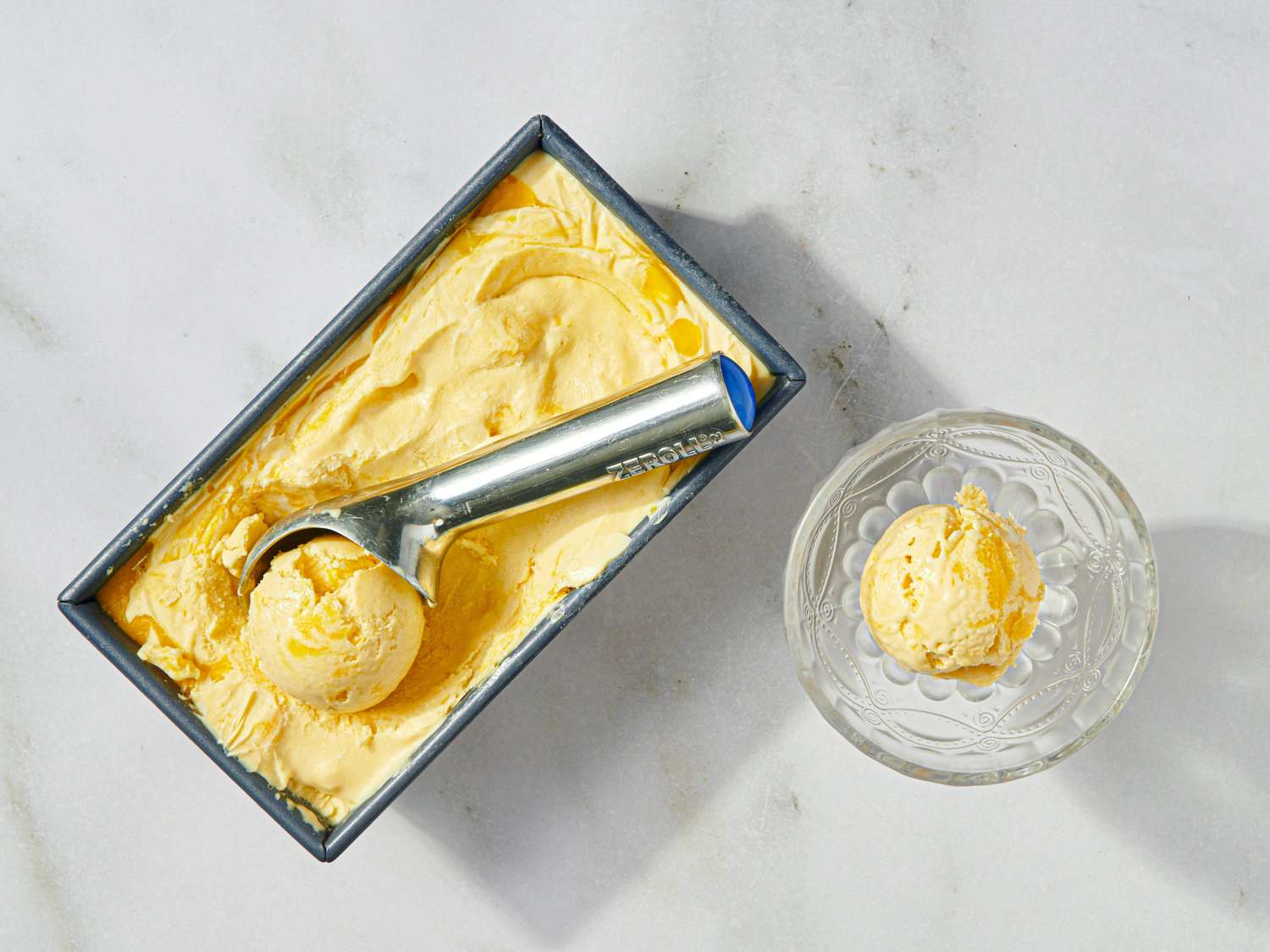 Mango ice cream in a rectangular tray with an ice cream scoop and a serving in a glass bowl