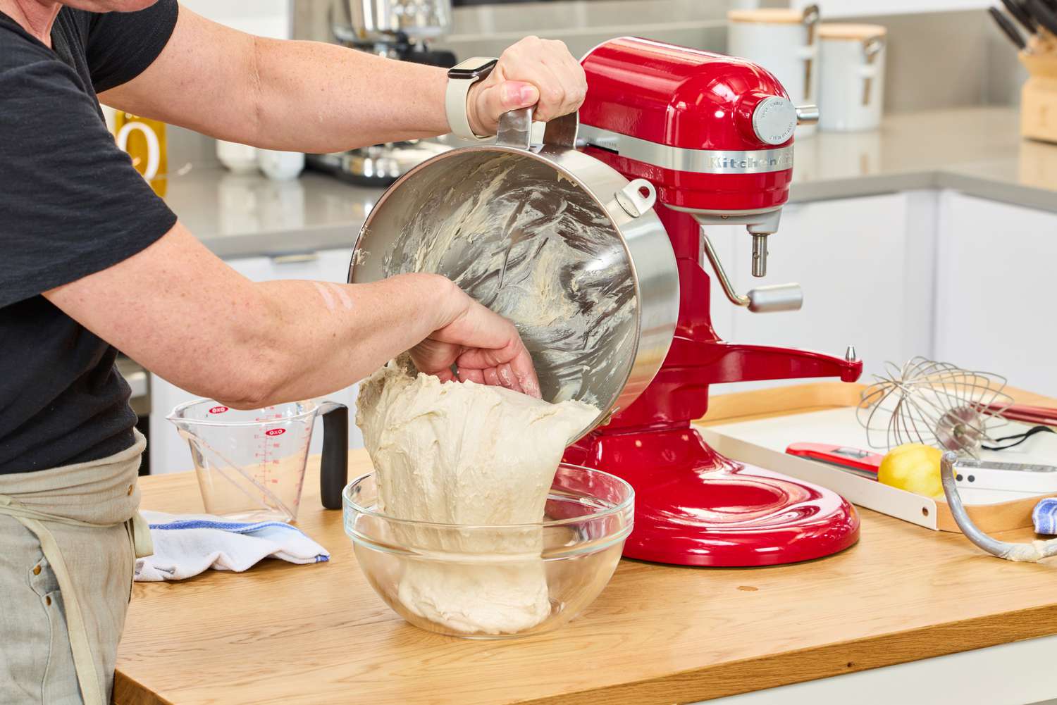 A person removing bread dough from a stand mixer's bowl into a mixing bowl.