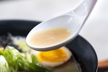 Closeup of a spoonful of chicken paitan broth. A bowl of ramen is visible in the blurred background.