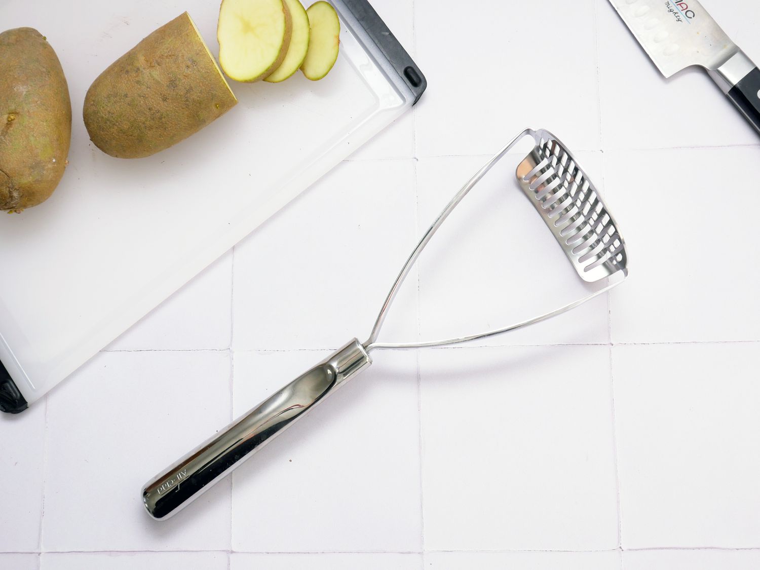 the all-clad potato masher on a white tile backdrop with a sliced potato 