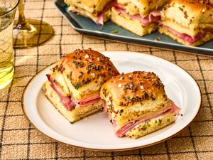 Ham and cheese sliders on a plate garnished with a poppy seed topping with additional sliders on a tray in the background