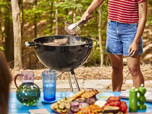 Person grilling on a barbecue grill in a wooded outdoor setting, food laid out on a table in the foreground
