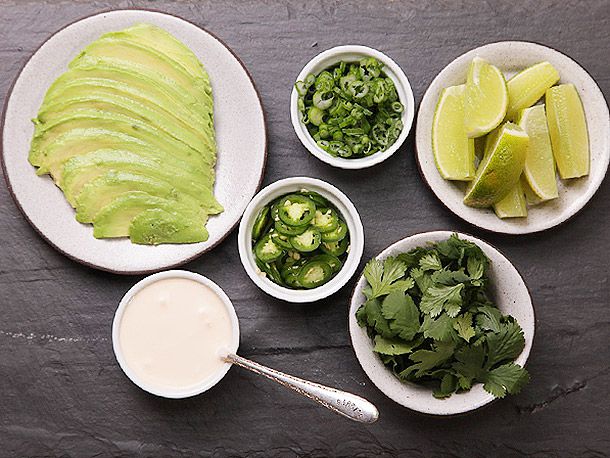 Black bean soup garnishes—sliced avocado, sliced scallions, sliced serrano peppers, crema, cilantro, and lime wedges—in white bowls on a black countertop.