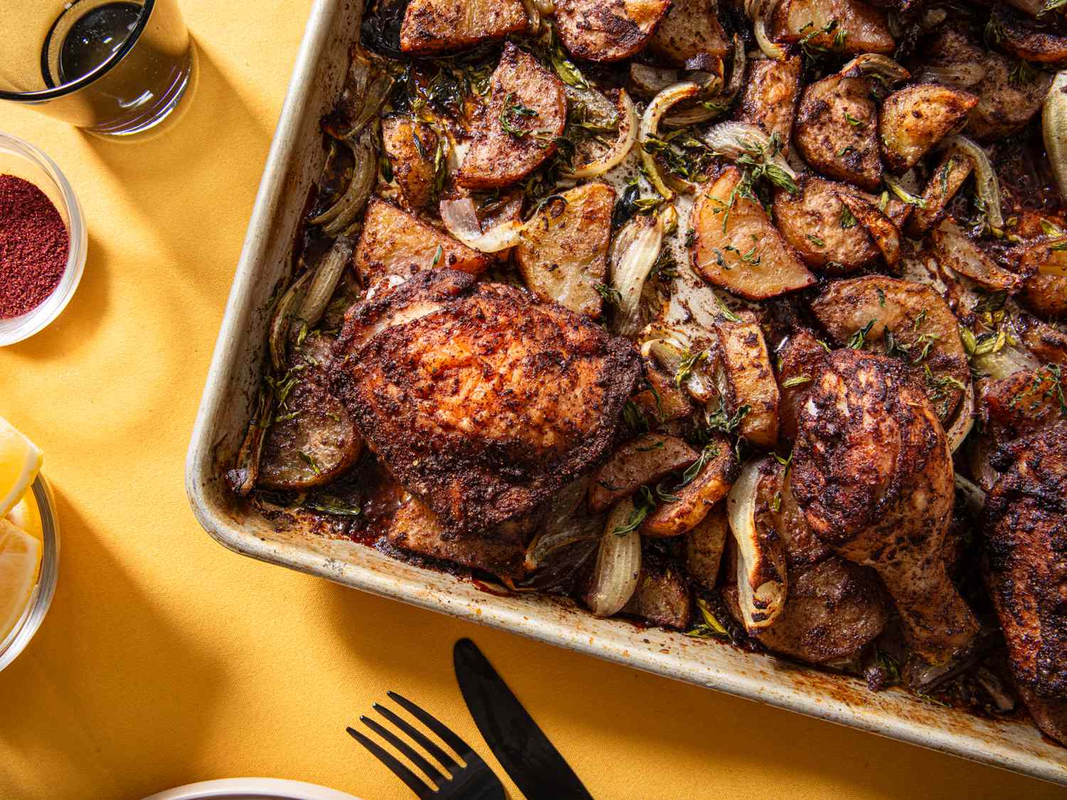 A sheet pan with seasoned chicken and roasted potato wedges on a yellow tablecloth served with sides and utensils visible