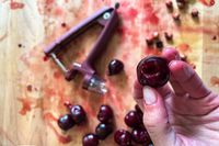 A closeup of a pitted cherry with a juice-stained cutting board and cherry pitter in the background