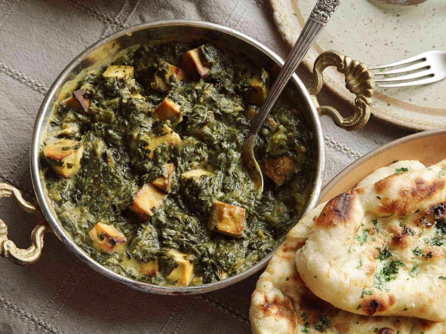 Overhead view of creamy vegan saag paneer in a serving vessel, flanked by a plate of naan.