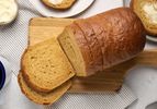 Sliced loaf of anadama bread on a wooden board, with two slices on plates, with grey striped napkin, mug of coffee, plate, butter, and breadknife behind it 