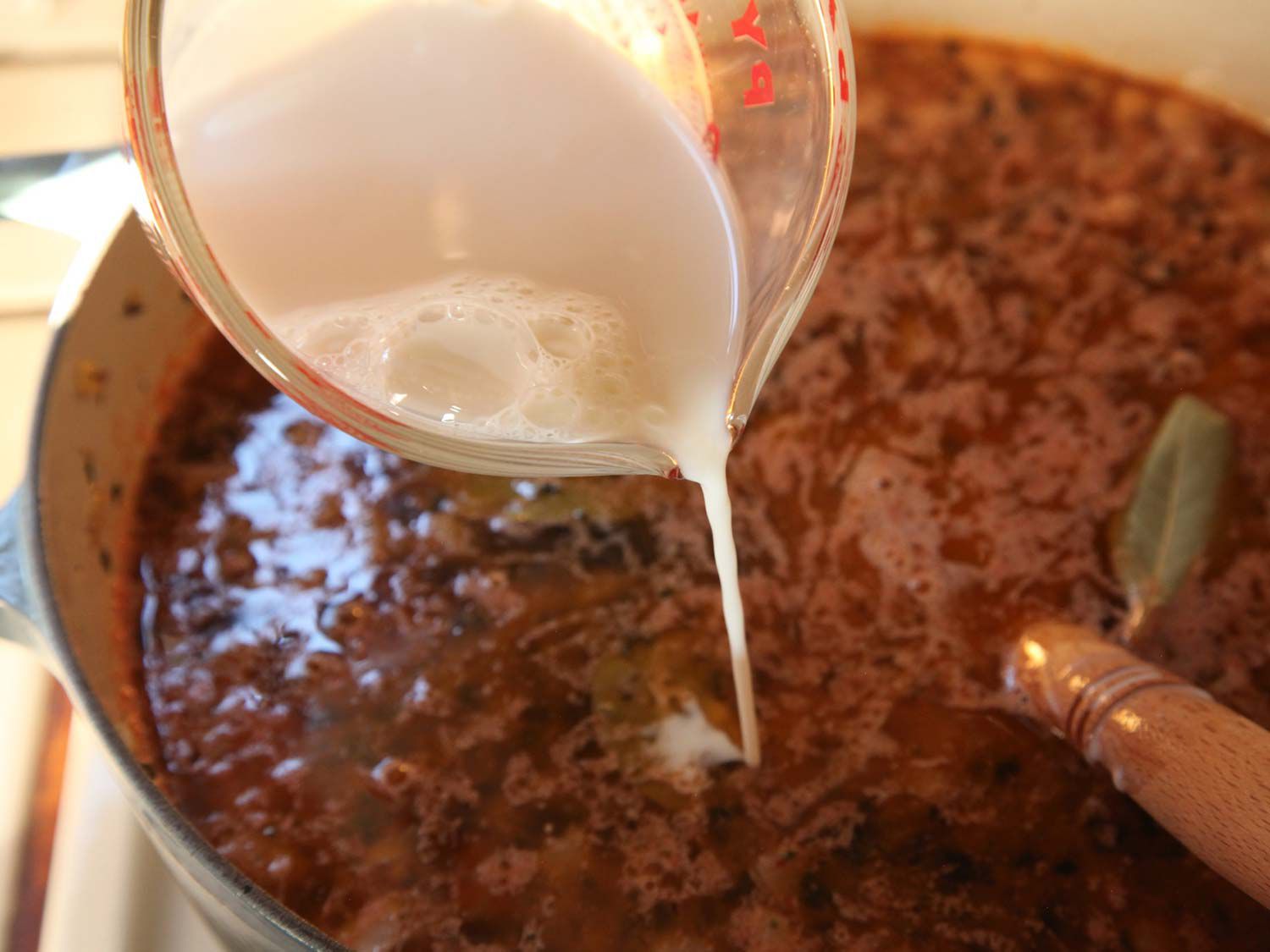 Milk in a measuring glass being poured into a meat sauce that is cooking in a Dutch oven.
