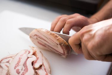 Knife cutting frozen pancetta into slices.